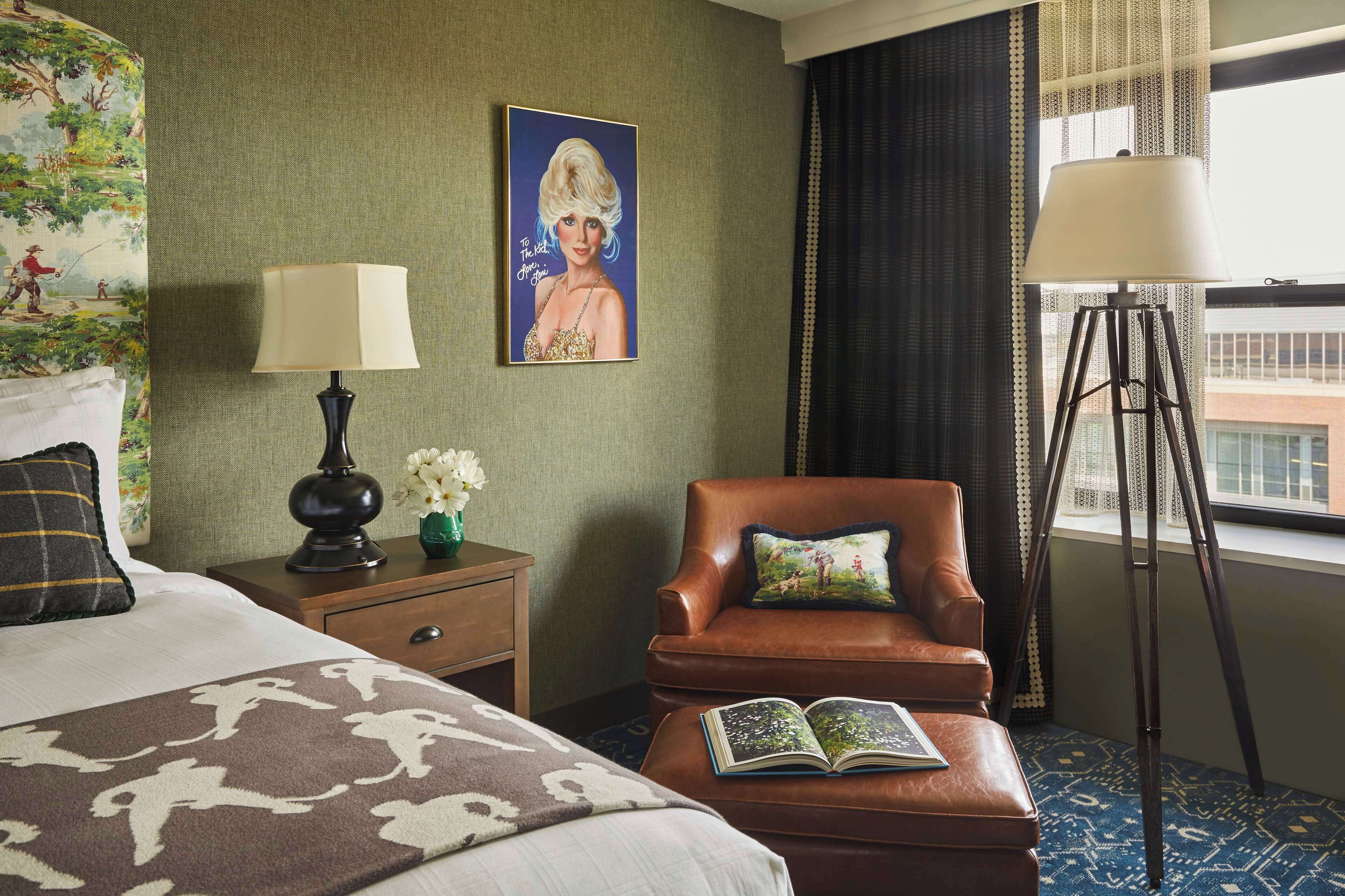 Habitación de hotel con cama, un sillón de cuero marrón, lámpara de lectura, un cuadro en la pared y una ventana con cortinas.