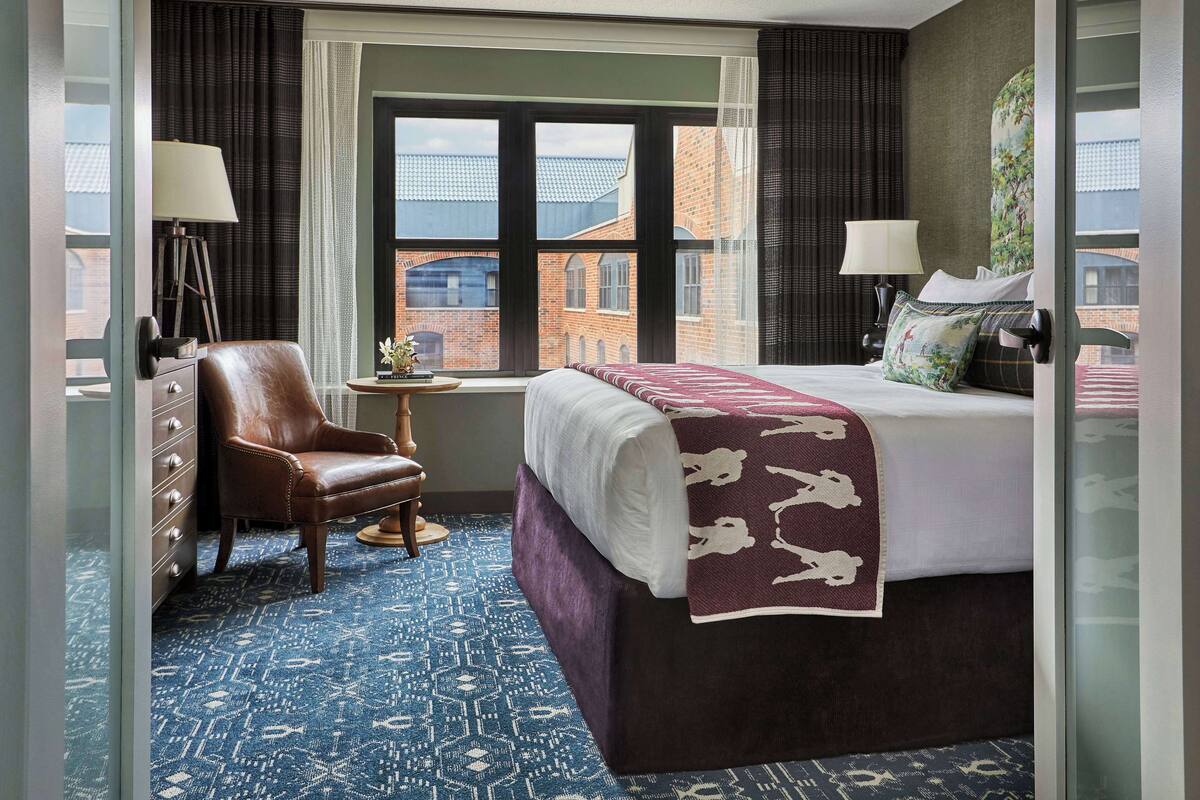 Ein Hotelzimmer mit einem großen Bett, einem Sessel, einem kleinen Tisch und Blick auf die Stadt durch ein Fenster.