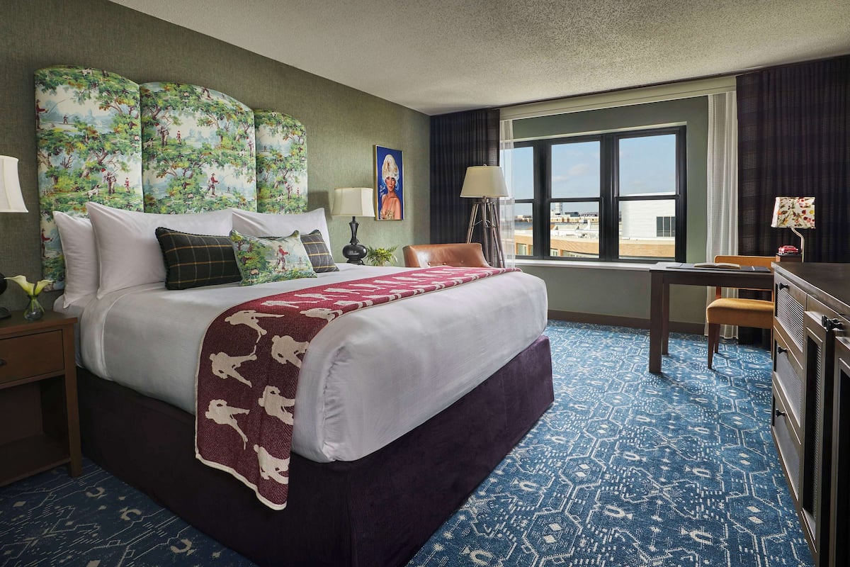 Ein Hotelzimmer mit einem großen Bett, einem Schreibtisch und Blick auf die Stadt durch das Fenster.