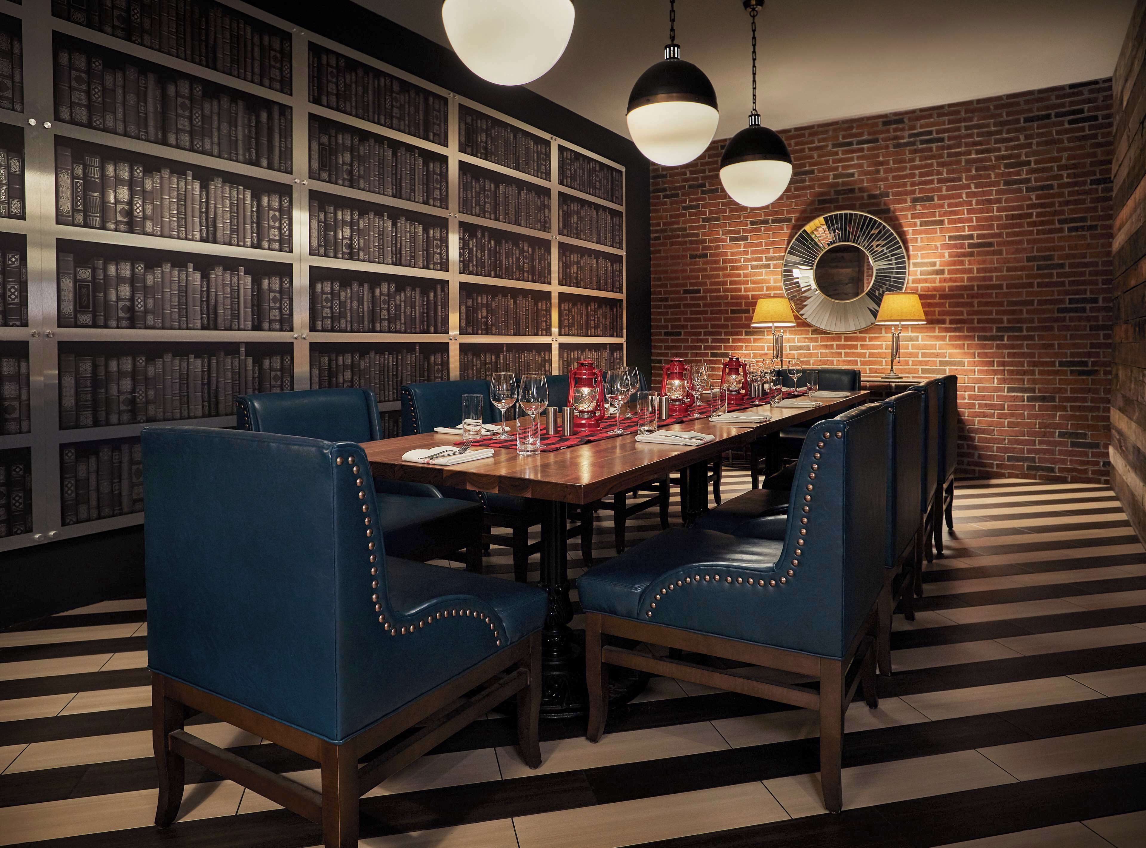 A dining area with a long table, blue upholstered chairs, and a brick wall with a bookshelf in the background.