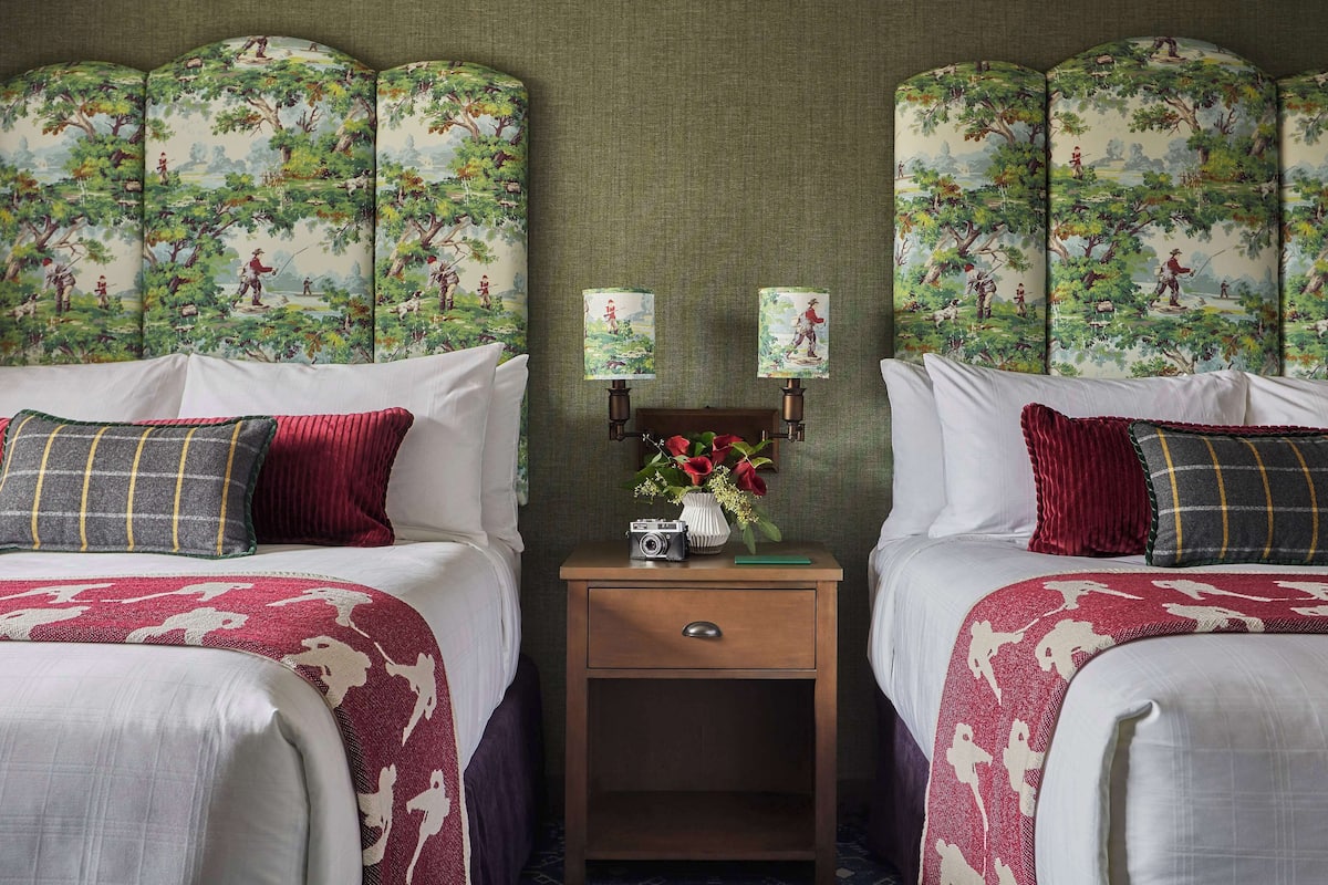 Ein Hotelzimmer mit zwei Betten, jeweils mit einer rot-weiß gemusterten Tagesdecke, einem kleinen Nachttisch mit Blumenvase und einer Zimmerpflanze.