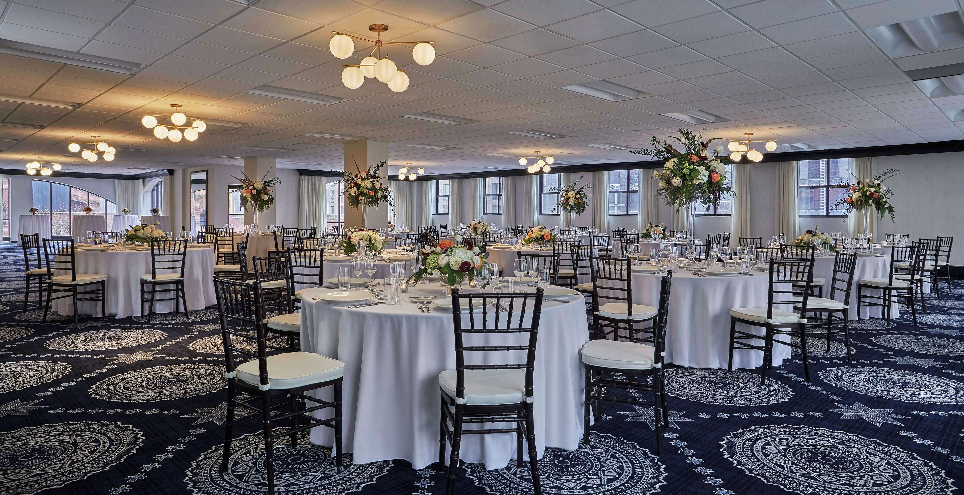 A large banquet hall with round tables, white tablecloths, and black chairs.