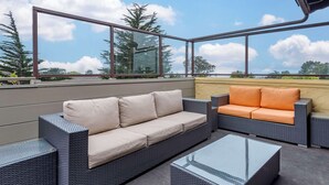 Een terras op het dak met rieten meubels, glazen tafels en een helder uitzicht op de lucht en bomen.