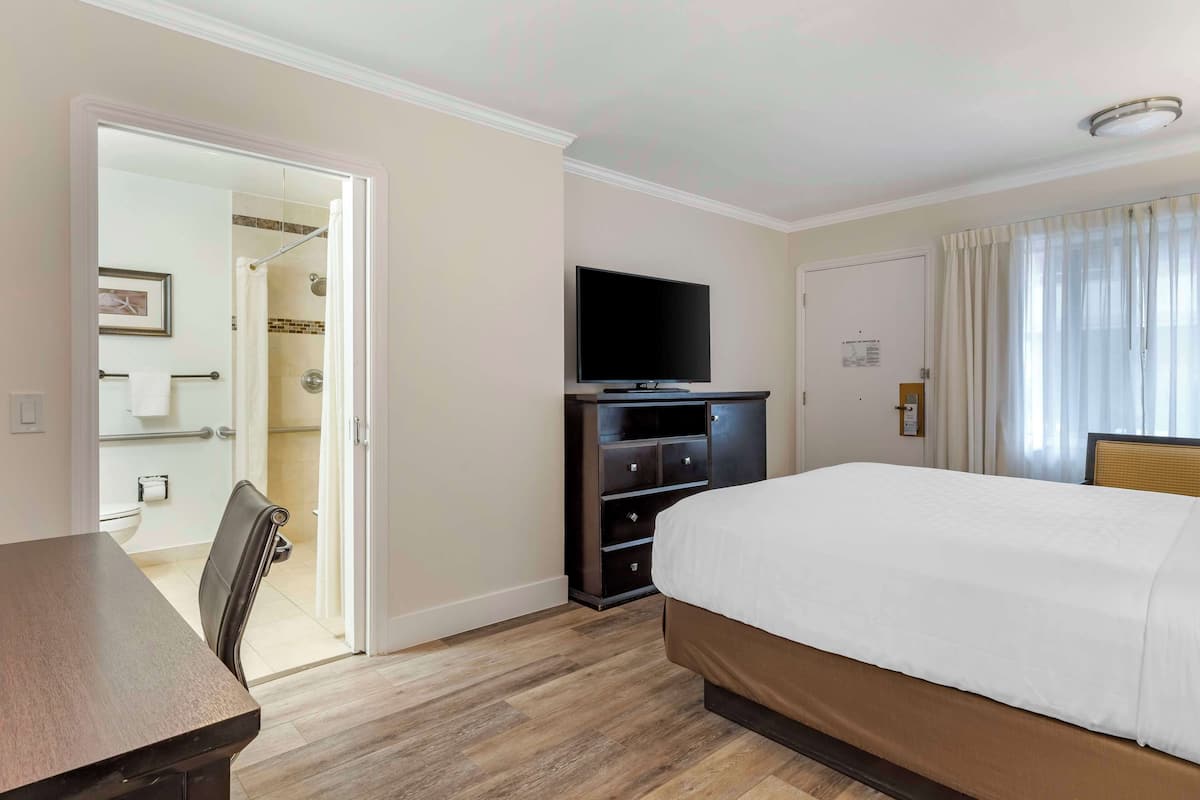 Ein Hotelzimmer mit Bett, Fernseher, Schreibtisch und Stuhl. Ein Badezimmer ist durch eine geöffnete Tür sichtbar.