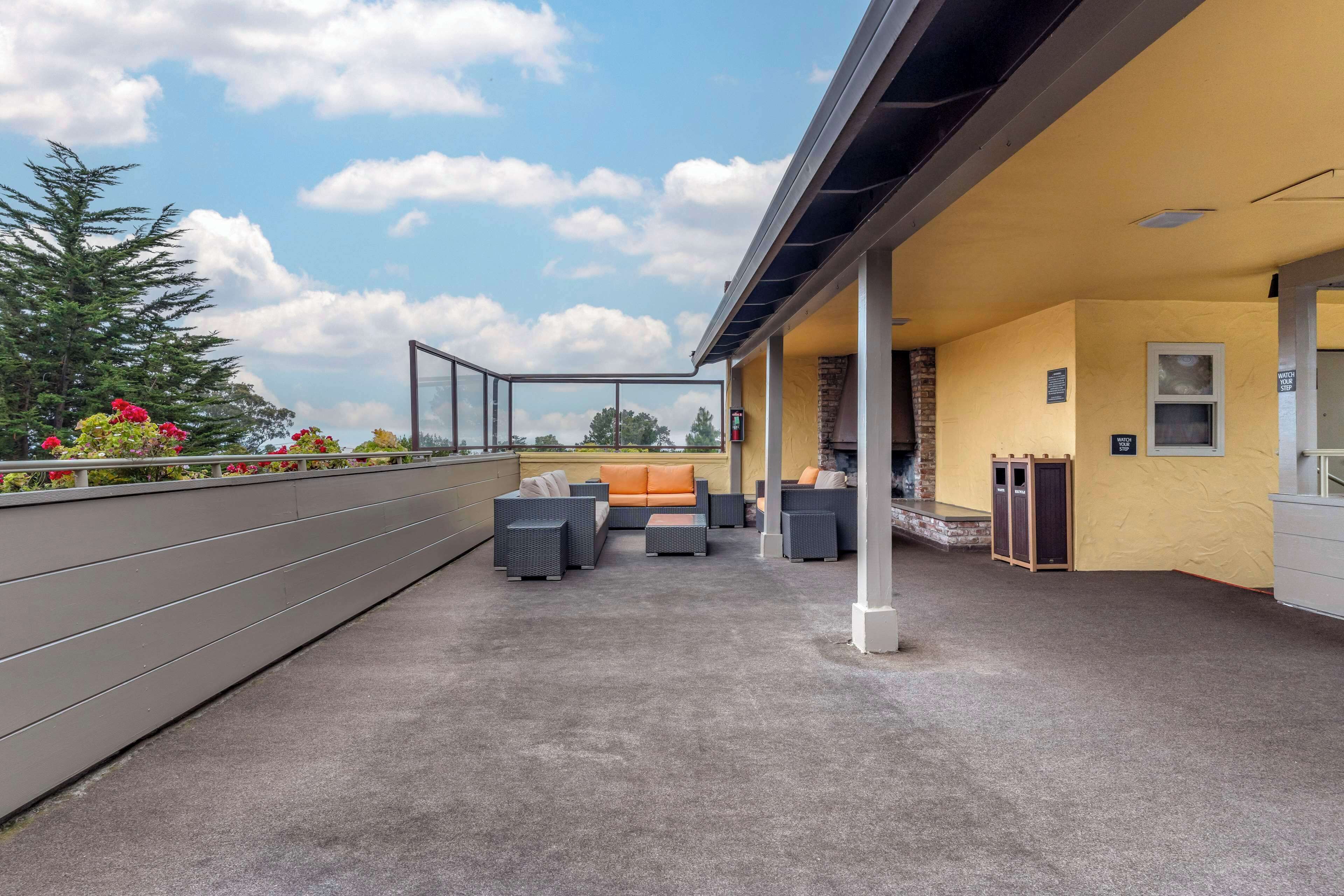 A covered outdoor seating area with sofas and chairs, a small kitchen area, and a view of trees and sky.