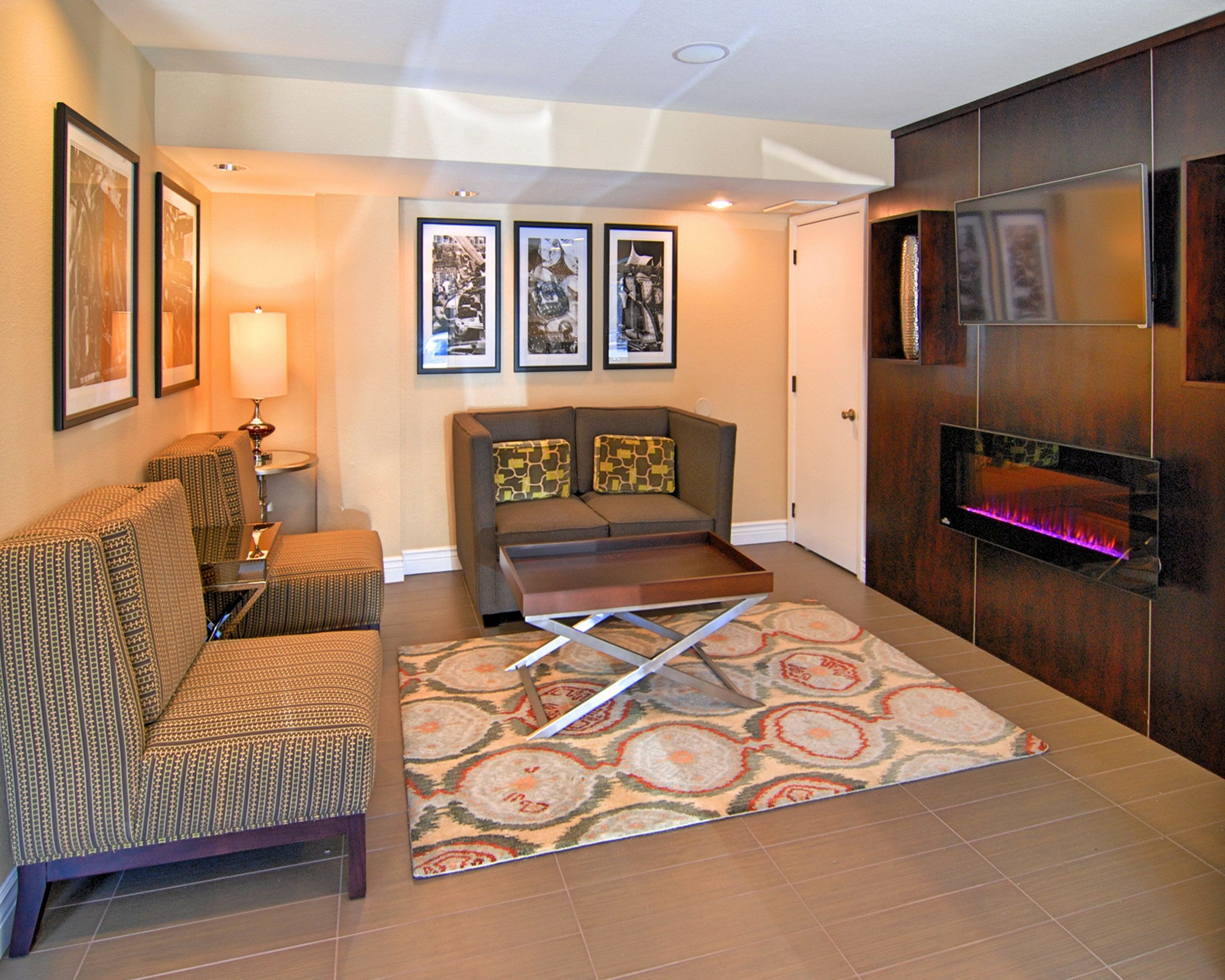 A hotel lobby with a sofa, armchairs, a patterned rug, framed pictures on the wall, and a fireplace with a pink light.