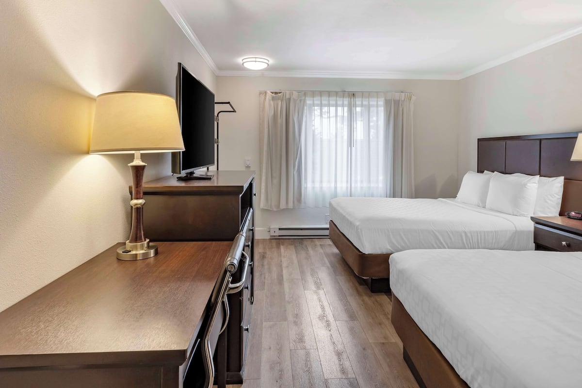Ein Hotelzimmer mit zwei Betten, einem Schreibtisch mit Lampe, einem Fernseher und einem Fenster mit Vorhängen.
