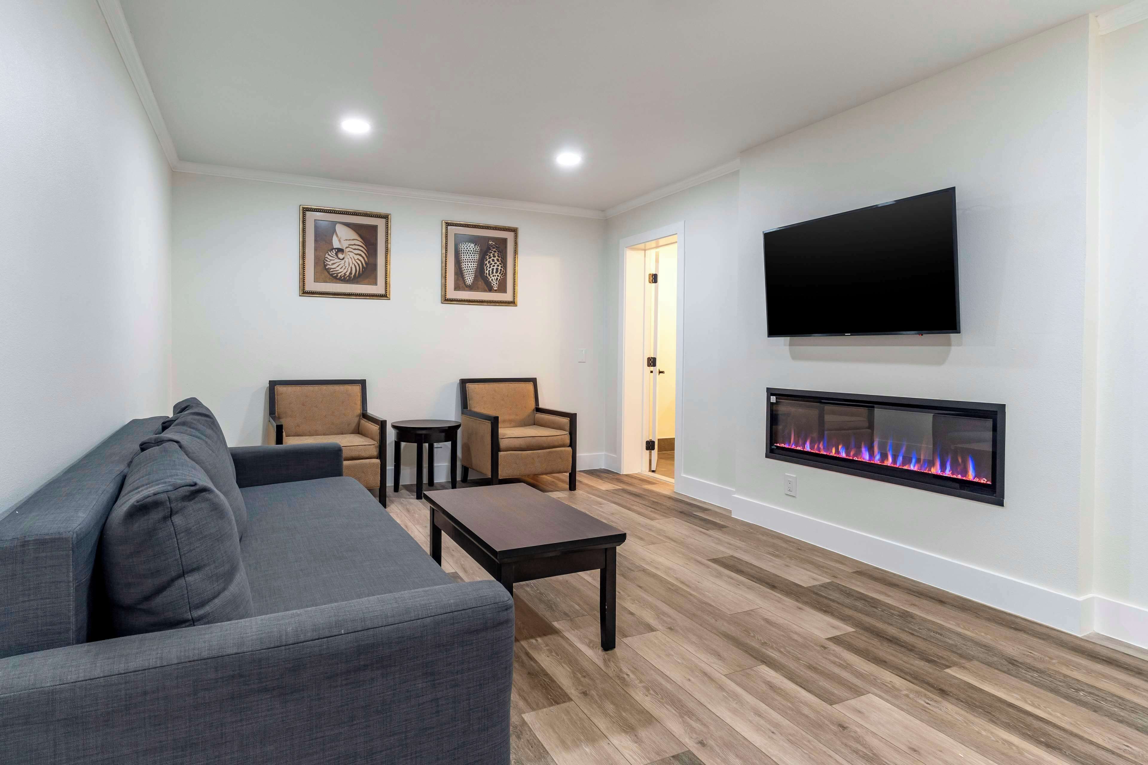 A living room with a gray sofa, two armchairs, a coffee table, a flat-screen TV mounted on the wall, and a fireplace with a visible fire.