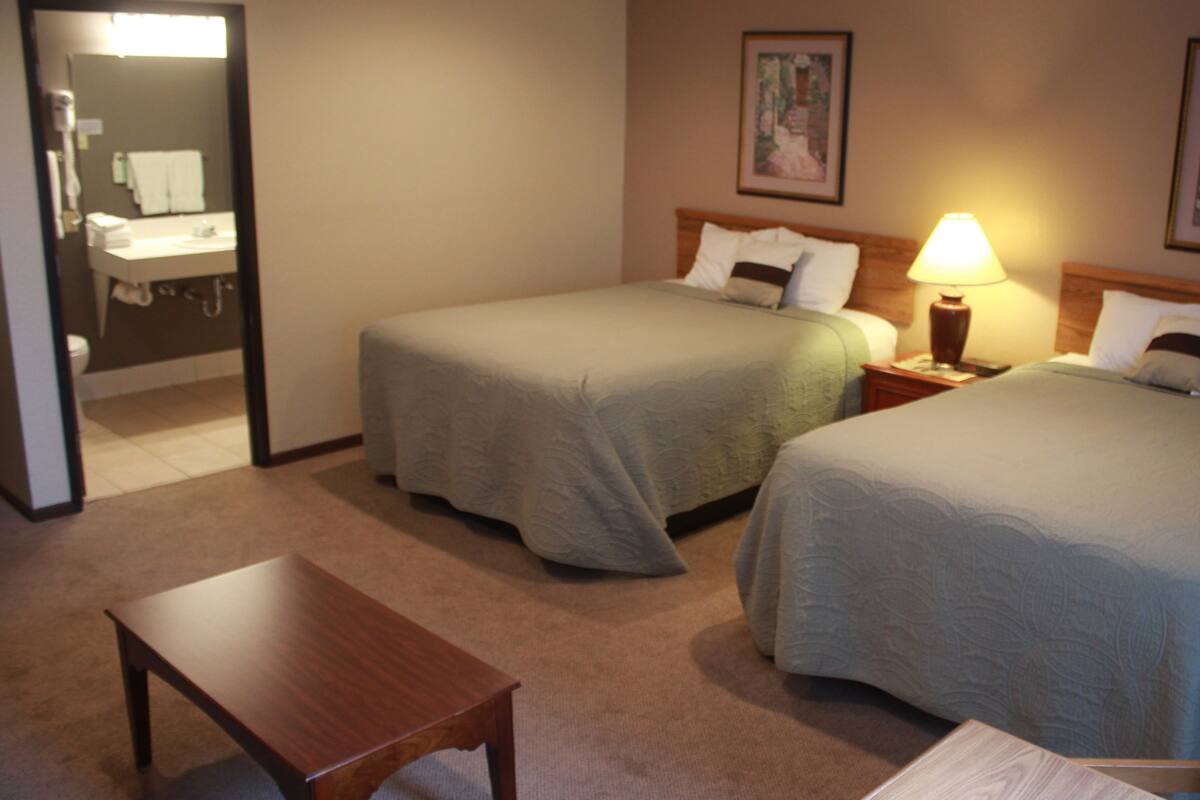 A hotel room with two beds, a wooden nightstand with a lamp, a small table, and a bathroom visible through an open door.