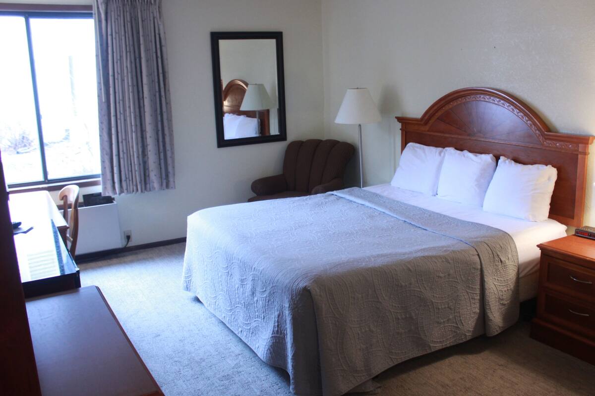 A hotel room with a large bed, a wooden headboard, a bedside table with a lamp, a chair, and a desk.