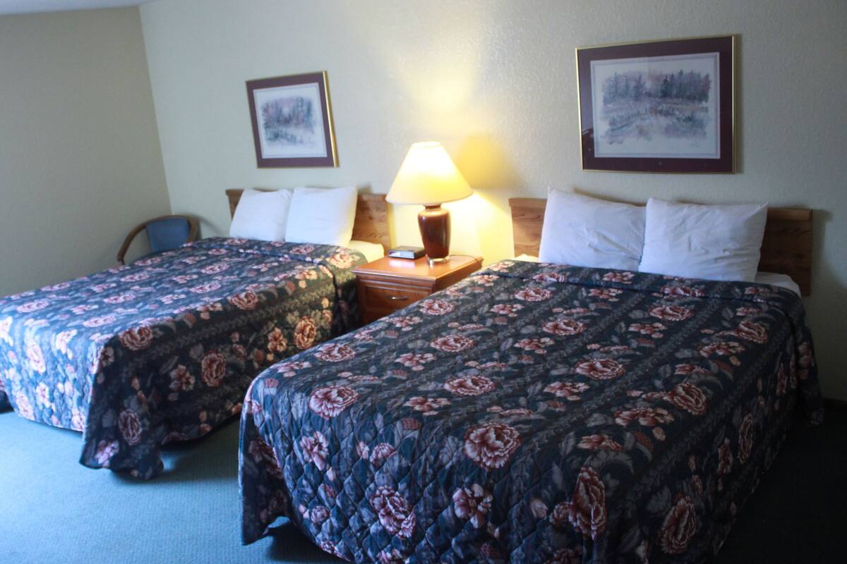 A hotel room with two beds, a nightstand with a lamp, a chair, and two framed pictures on the wall.