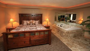 A bedroom with a large bed, a wooden headboard, two bedside lamps, a large mirror, and a separate bathroom with a bathtub. - Shawano Four Seasons Hotel (Shawano)