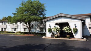 A hotel entrance with a white facade, large windows, and a tree in front. There are flower arrangements on the steps. - Shawano Four Seasons Hotel (Shawano)