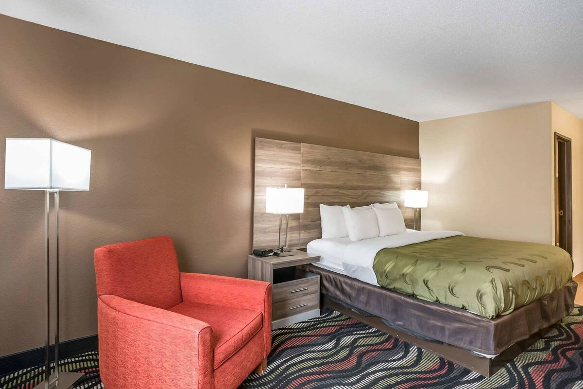 Una habitación de hotel con una cama, una silla roja, una mesita de noche, una lámpara y una alfombra estampada.
