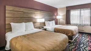 A hotel room with two beds, a large wooden headboard, a window with curtains, and a patterned rug. - Quality Inn Cedar Rapids South (Cedar Rapids)