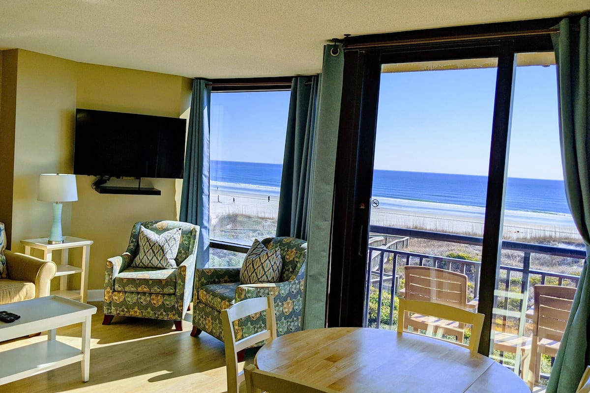 Ein Zimmer mit einer Couch, einem Sessel und einem Tisch, einem an der Wand befestigten Fernseher und einem Balkon mit Blick auf den Strand.