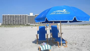 Un parasol de plage arborant le logo de « Shell Islands Resort », deux chaises de plage bleues et une petite chaise en bois avec une serviette à rayures.
