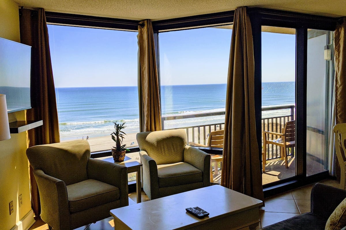 Ein Zimmer mit Blick auf den Strand, inklusive Fernseher, zwei Sesseln, einem Tisch und einer Fernbedienung.