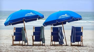 Cuatro sillas de playa con toallas azules, dos sombrillas azules con la marca 'Shell Island Resort', y una playa de arena con el océano al fondo.