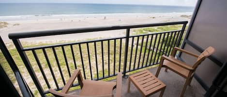 Un balcón con dos sillas de madera y una mesa pequeña, con vista a una playa y una vista despejada del océano.