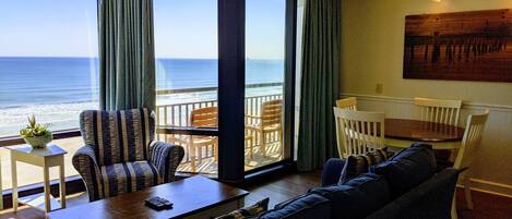 A room with a view of the ocean, a sofa, a dining table with chairs, and a small table with a plant.