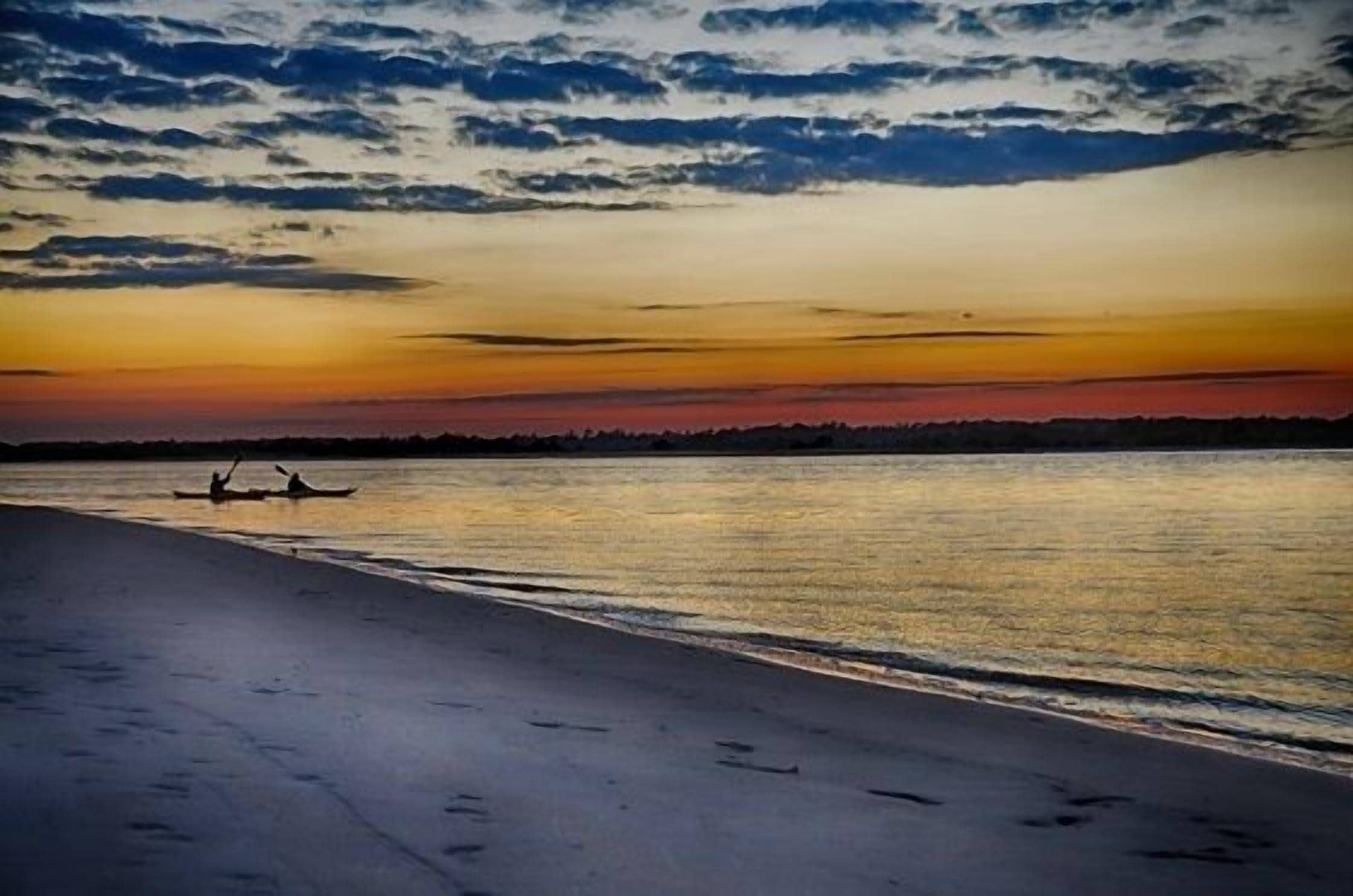 Une scène de plage au coucher du soleil, avec une eau calme et deux personnes qui pagayent dans un canoë.