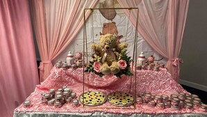A table with pink floral tablecloth, jars of food, and a teddy bear centerpiece.