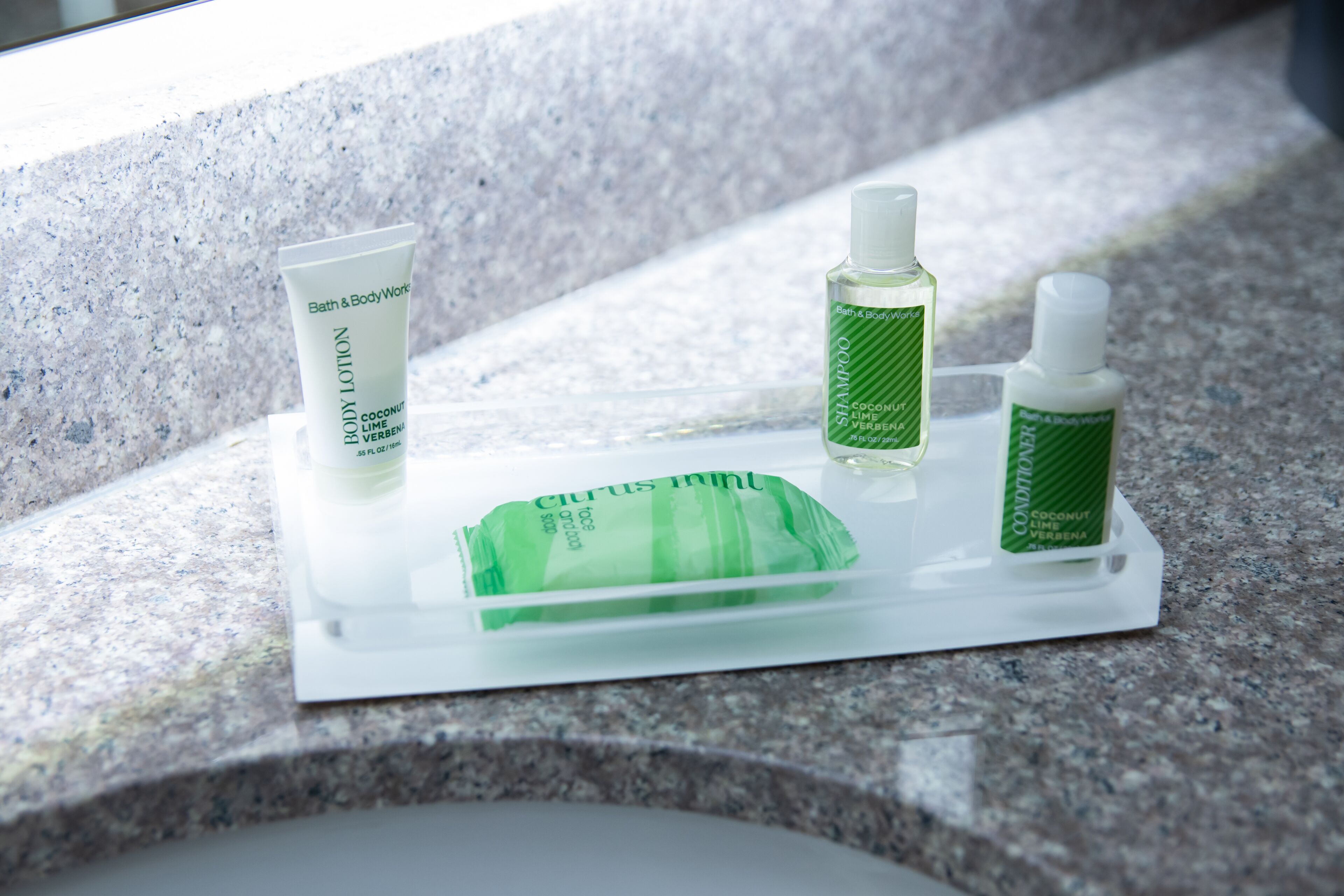 A bathroom counter with a white tray holding various toiletries including a body lotion, shampoo, conditioner, and a green bar of soap.