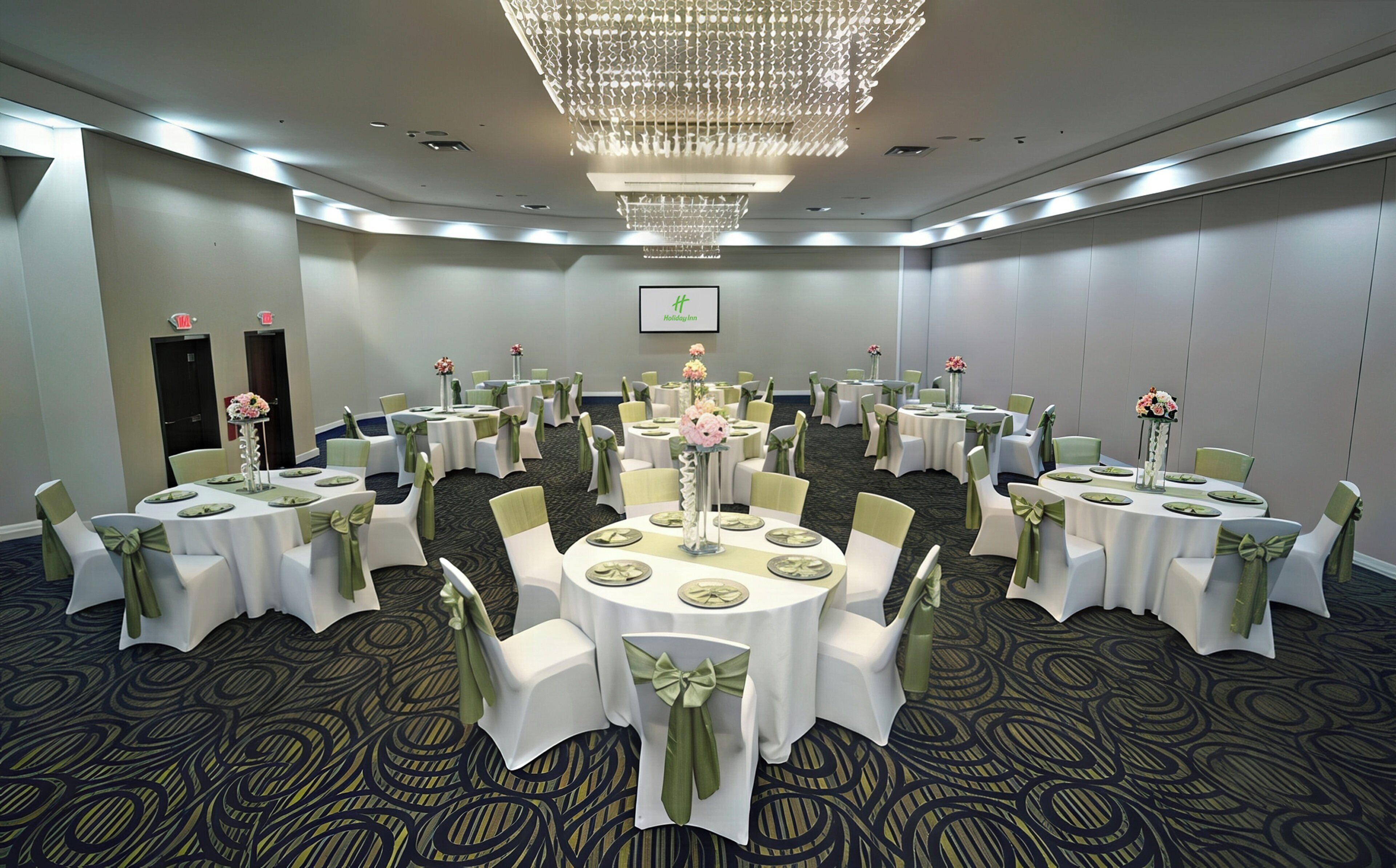 A conference room with multiple round tables, each with white tablecloths and green chair covers, arranged in a U-shape.
