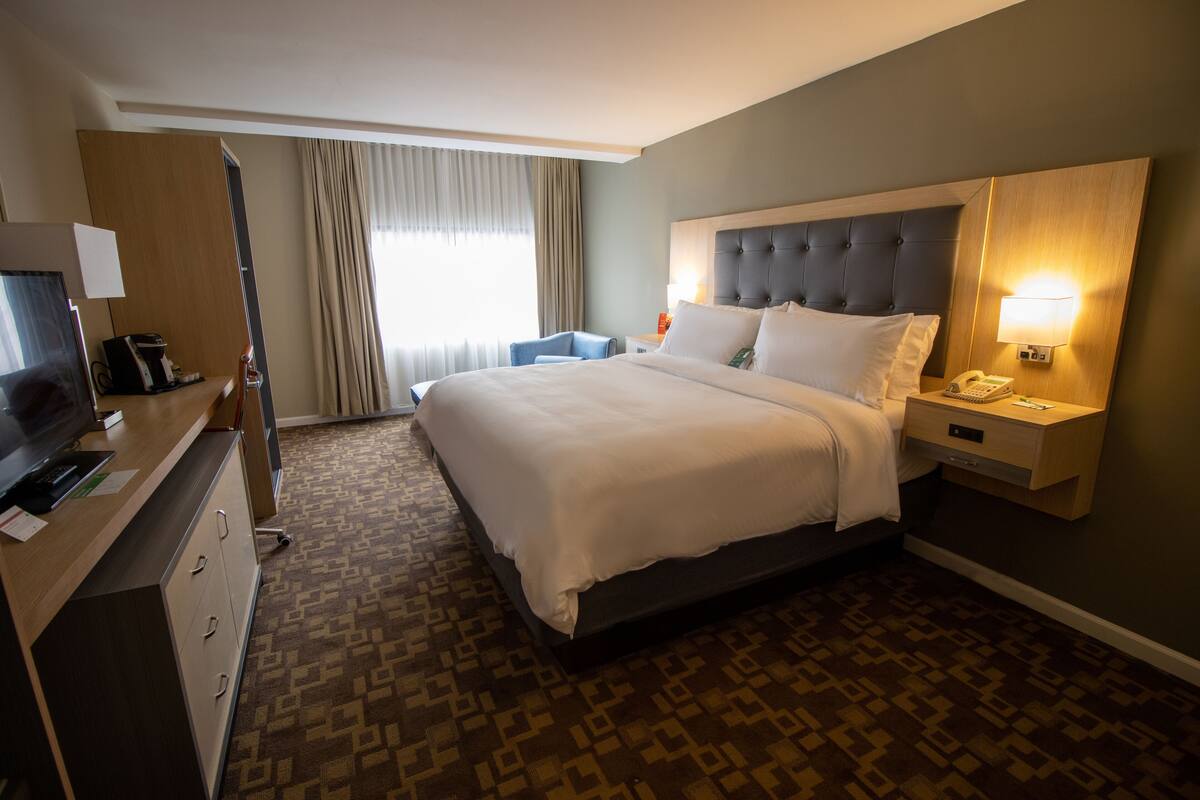 A hotel room with a large bed, a desk with a computer, a chair, a TV, and a window with curtains.