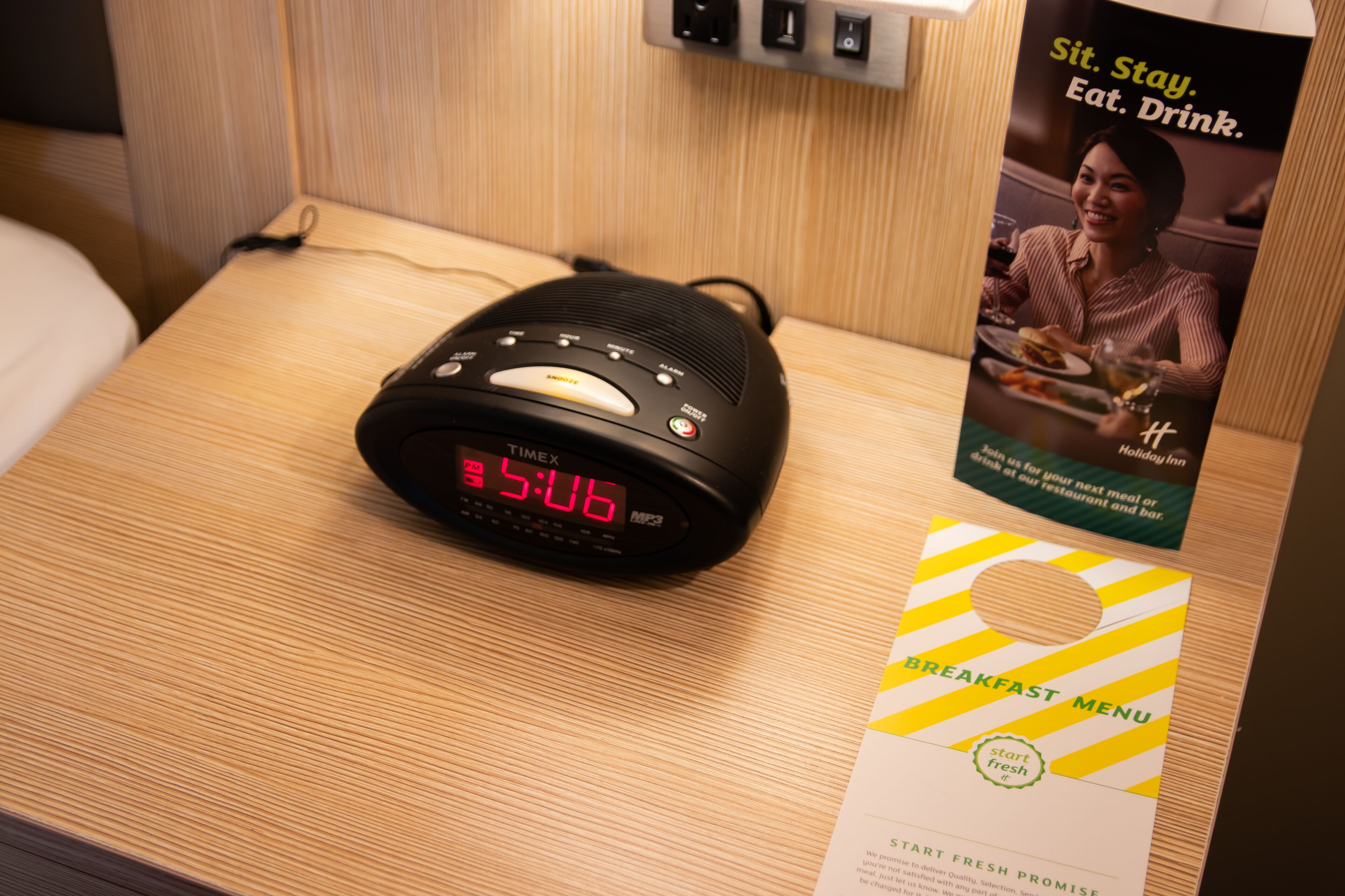 A digital clock displaying 3:06, a breakfast menu, and a poster with the text "Sit. Stay. Eat. Drink."