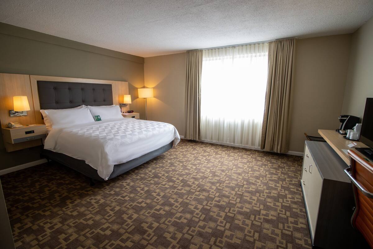 A hotel room with a large bed, two bedside tables with lamps, a desk with a chair, and a large window with curtains.