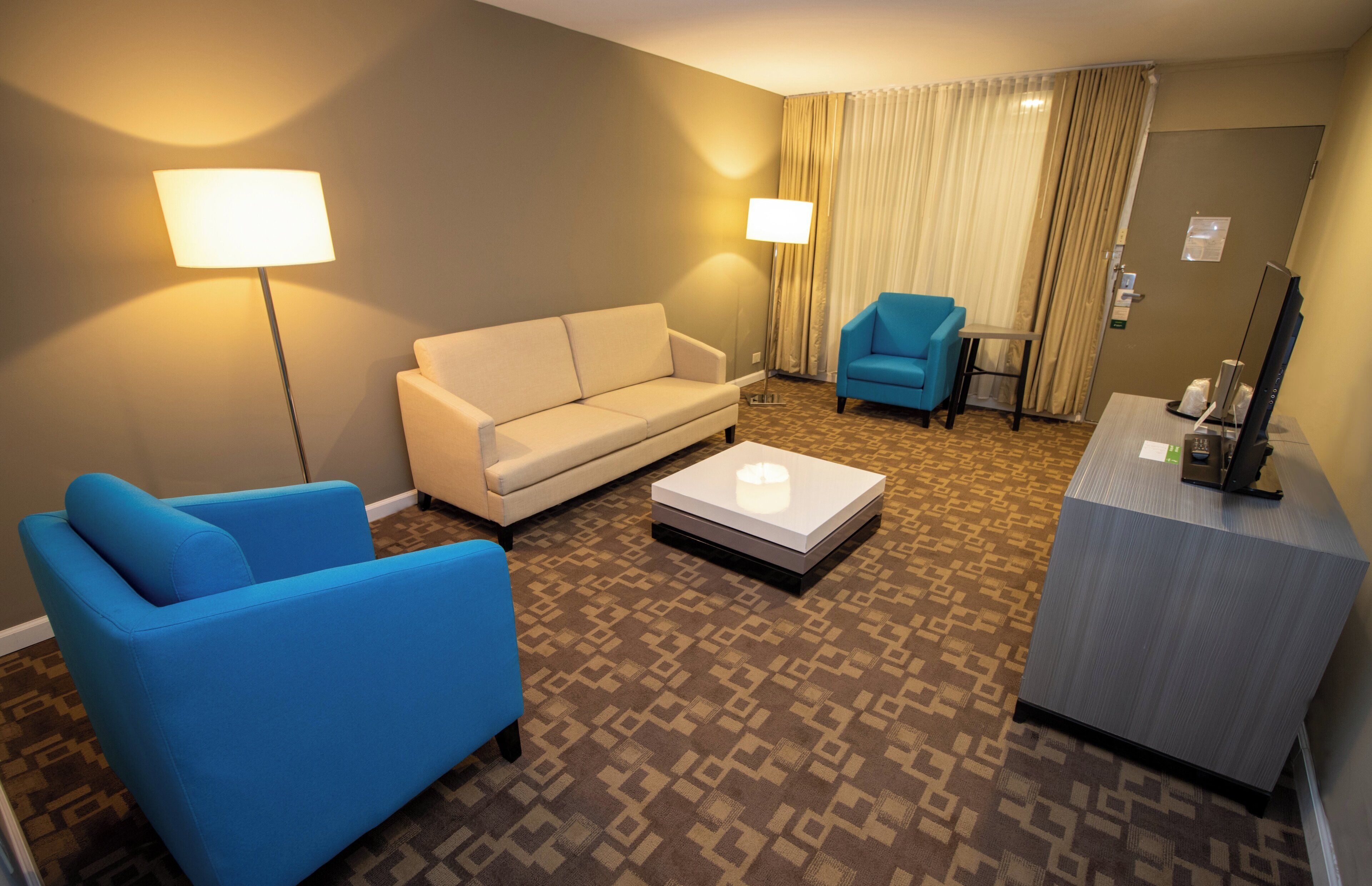Una habitación de hotel con sofá, un sillón azul, una mesita, un escritorio con computadora y una alfombra estampada.