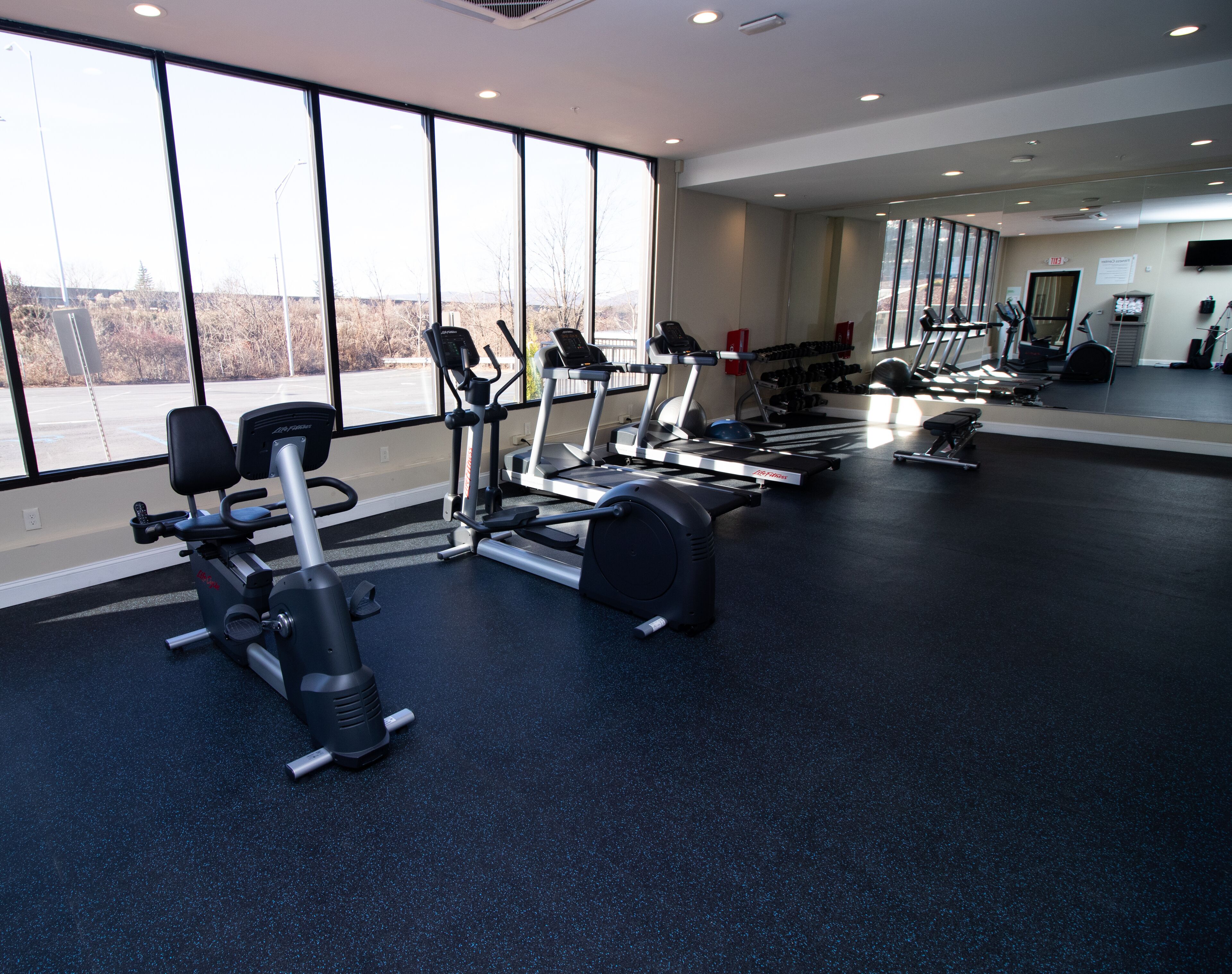A gym with various exercise equipment, large windows, and a mirror on the wall.