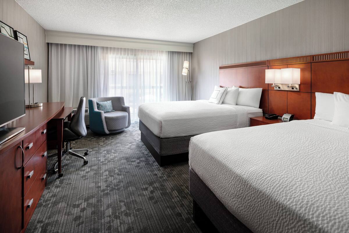 A hotel room with two beds, a desk, a chair, and a TV.