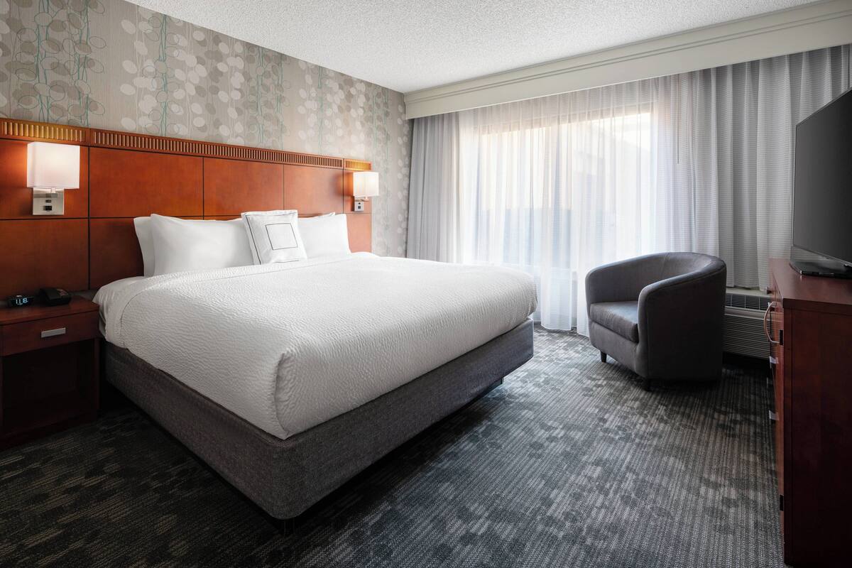 A hotel room with a large bed, a chair, a TV, and a desk with a lamp.