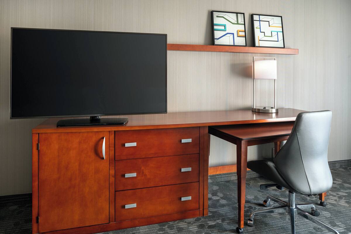 A hotel room with a wooden desk, a flat-screen TV, a chair, and a lamp.