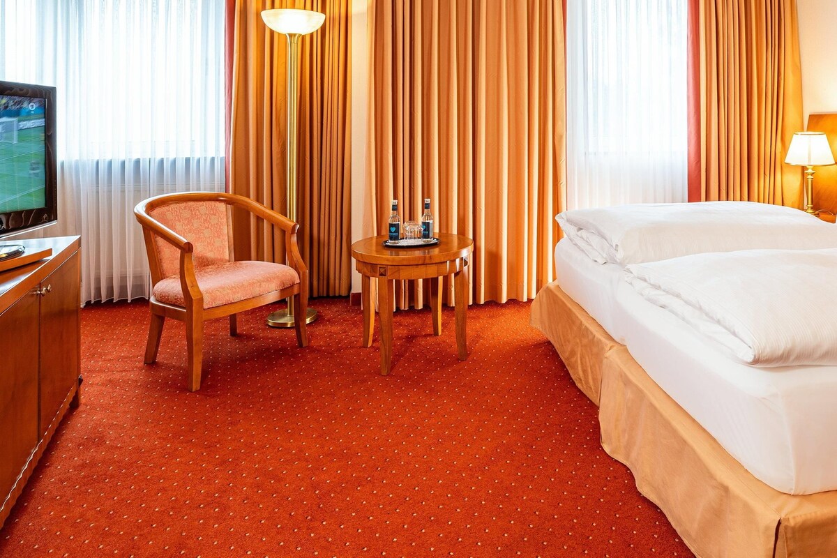 Una habitación de hotel con cama, una silla, una mesita con bandeja, televisión y cortinas.
