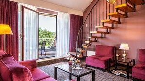 A modern hotel room with a red sofa, a glass coffee table, a red armchair, a small table with a lamp, and a spiral staircase.