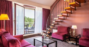 Una habitación de hotel moderna con un sofá rojo, una mesa de centro de vidrio, un sillón rojo, una pequeña mesa con una lámpara y una escalera de caracol.