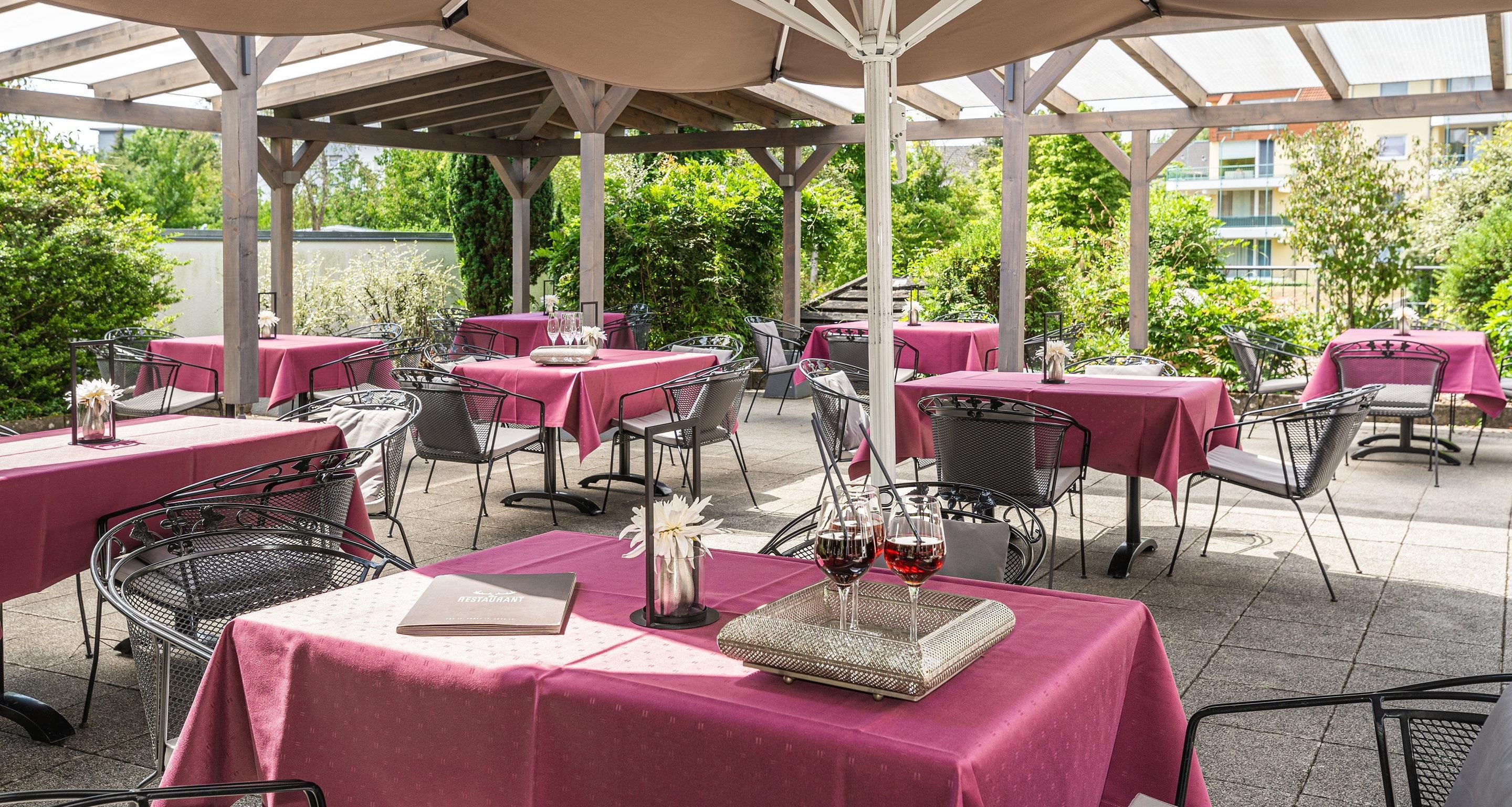 Eine Terrassenanlage im Freien mit Tischen, die mit rosa Tischdecken bedeckt sind, Metallstühlen und einer Pergola.