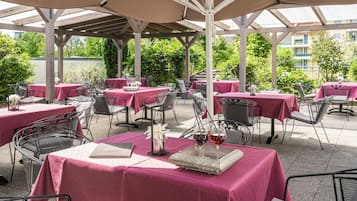 Eine Terrassenanlage im Freien mit Tischen, die mit rosa Tischdecken bedeckt sind, Metallstühlen und einer Pergola.