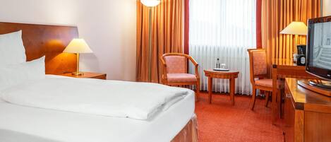 Habitación de hotel con cama, dos sillas, una mesita con lámpara, televisor y ventana con cortinas.
