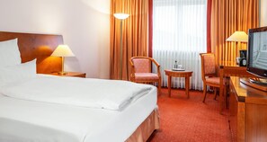 Habitación de hotel con cama, dos sillas, una mesita con lámpara, televisor y ventana con cortinas.