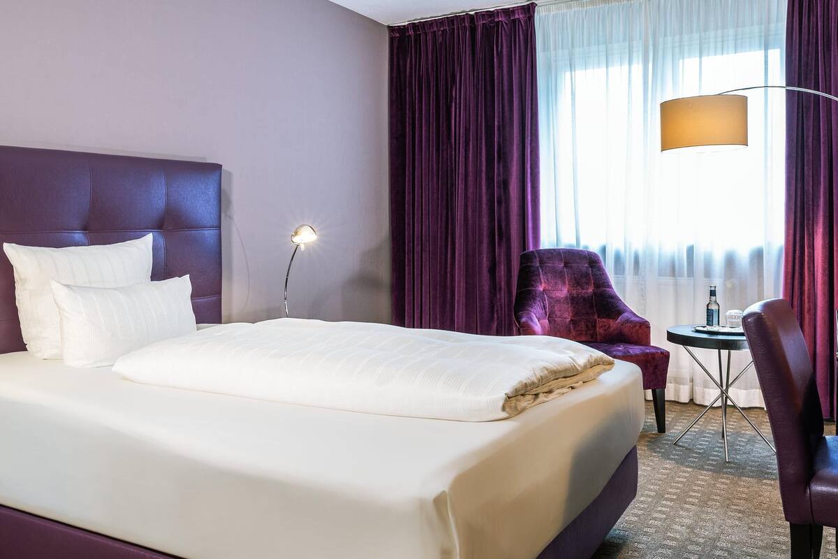A hotel room with a large bed, a desk with a lamp, a chair, and a view of the outside through curtains.