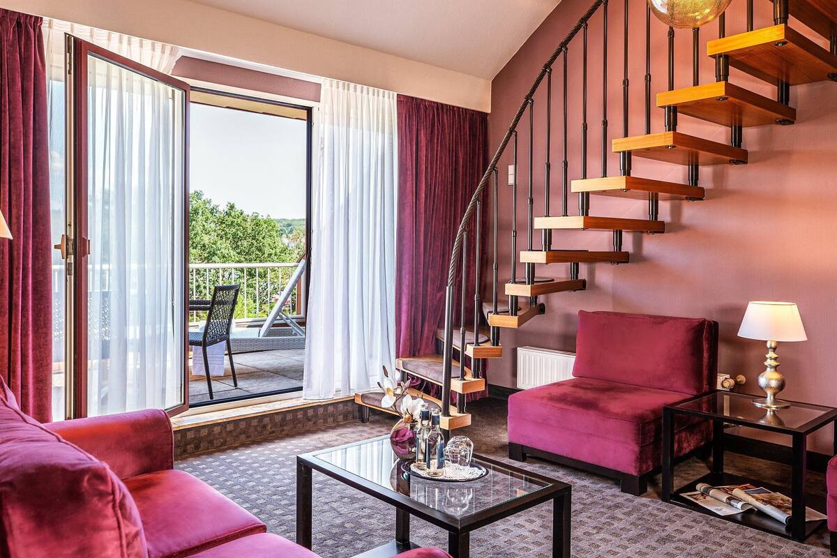 A modern hotel room with a red sofa, a glass coffee table, a red armchair, a small table with a lamp, and a spiral staircase.