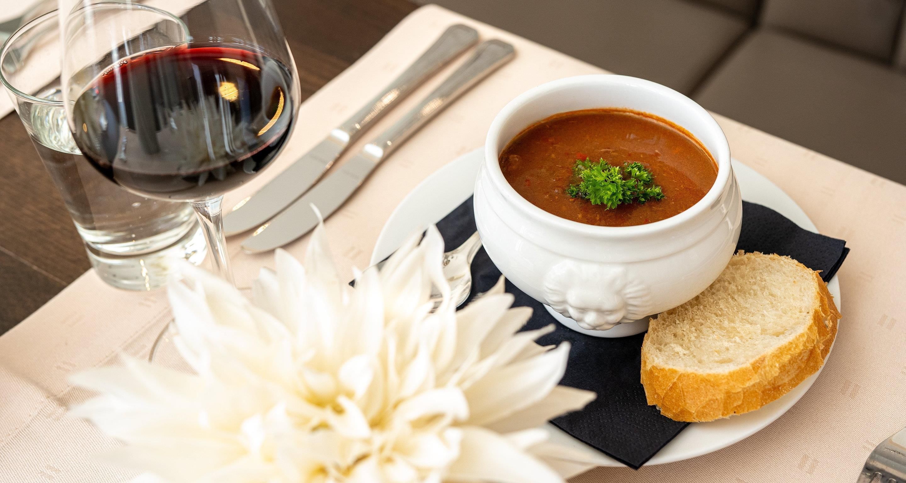 Une table à manger avec un bol de soupe, un verre de vin rouge, un couteau, un morceau de pain et une fleur blanche.