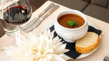 Una mesa de comedor con un tazón de sopa, una copa de vino tinto, un cuchillo, un trozo de pan y una flor blanca.