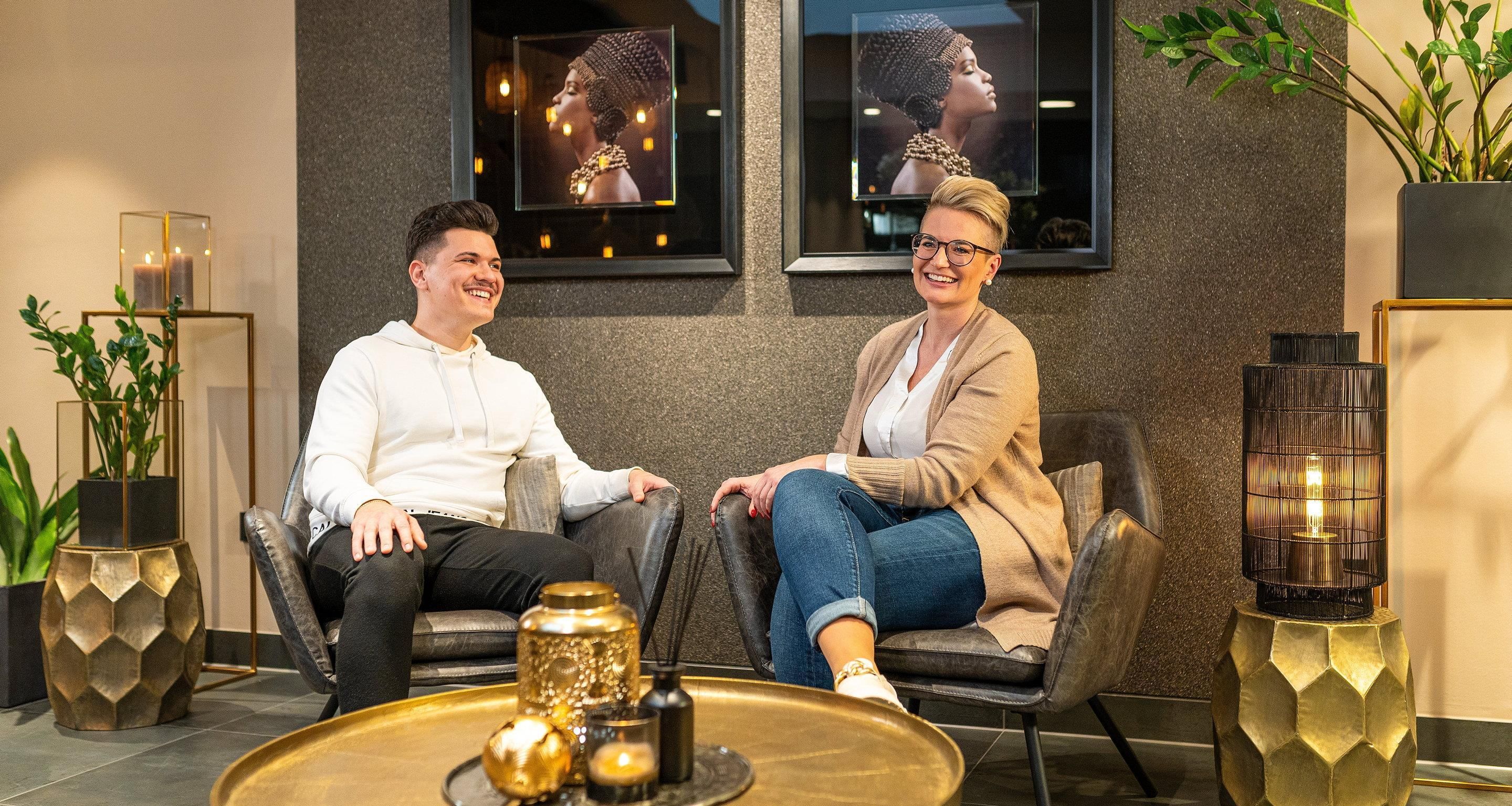 Two people seated on modern chairs in a well-lit room with a decorative table, framed artwork on the wall, and potted plants.
