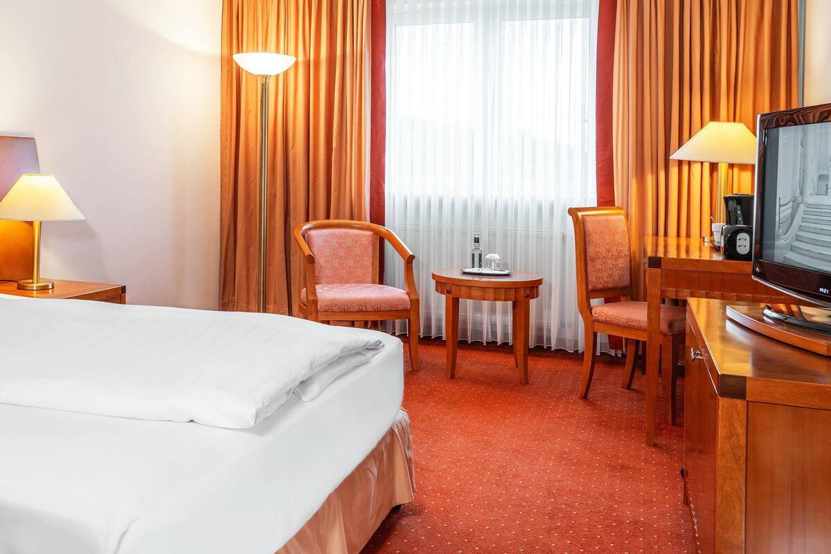 A hotel room with a bed, two chairs, a small table with a lamp, a television, and a window with curtains.